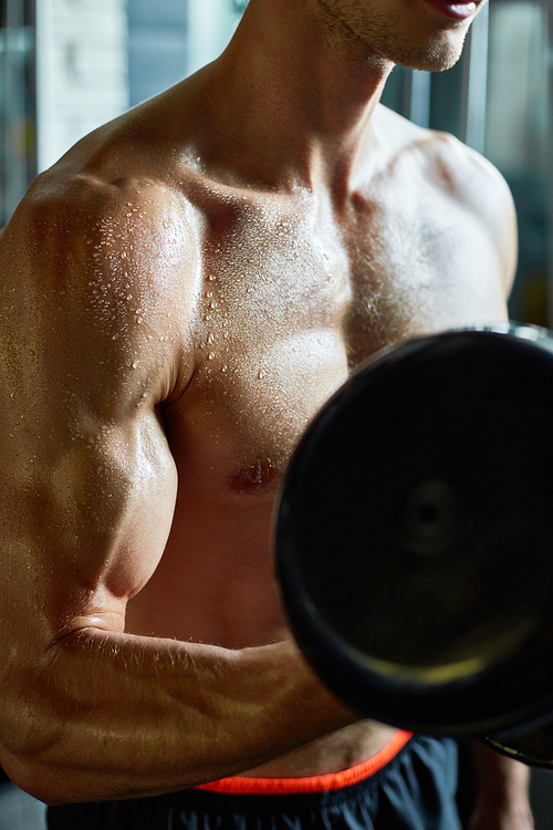 Close-up shot of sweaty muscular man pumping up muscles with dumbbell while having intensive training at gym