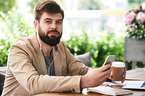 Portrait of modern bearded businessman using smartphone and  enjoying coffee break in outdoor cafe