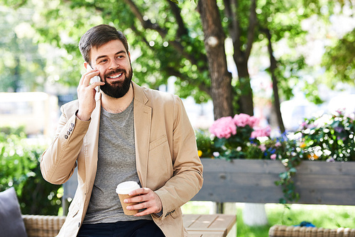 Portrait of young bearded businessman speaking by phone and smiling while enjoying coffee break outdoors