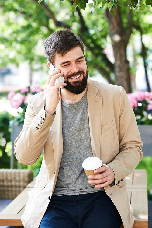 Portrait of modern bearded man speaking by phone and smiling while enjoying coffee break outdoors