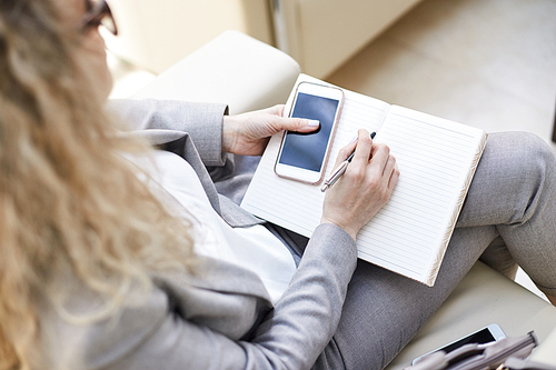 Profile view of unrecognizable curly businesswoman sitting on cozy sofa at office lobby, thinking over promising project and taking necessary notes, close-up shot