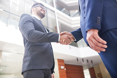 Confident business partners shaking hands after successful completion of negotiations, interior of modern office lobby on background, low angle view