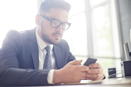 Waist-up portrait of confident bearded entrepreneur in classical suit texting with colleague on smartphone while sitting at office desk, blurred background