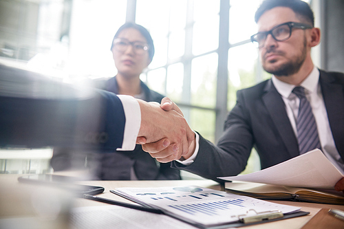 Confident bearded entrepreneur wearing elegant suit sitting at boardroom table and greeting business partner with firm handshake, pretty Asian assistant manager next to him