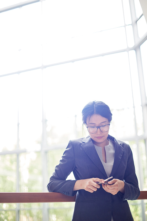 Attractive Asian entrepreneur in eyeglasses holding smartphone in hands while standing at spacious office lobby, glass wall on background