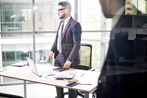 Portrait of pensive bearded entrepreneur looking out panoramic window while standing at office desk, view through glass wall