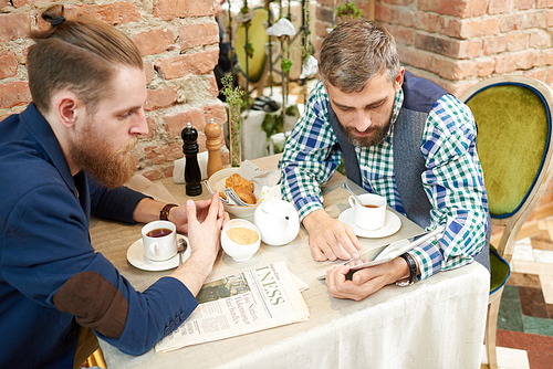 Gathering of best friends: bearded men watching video on digital tablet while enjoying fragrant coffee and pastries, interior of stylish coffeehouse on background