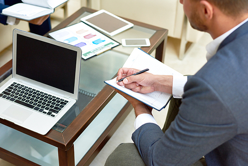 High angle closeup of successful businessman writing work notes in notebook sitting by coffee table with laptop during meeting, copy space