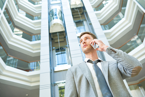 Low angle view of confident young businessman talking to his colleague on smartphone while standing at spacious office lobby, waist-up portrait shot