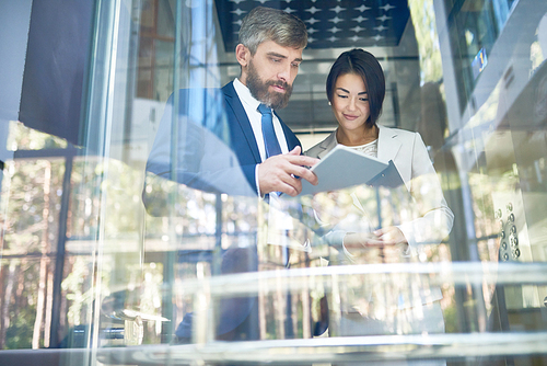 Pretty young manager and her middle-aged bearded colleague standing at panoramic window of office lobby and analyzing financial figures with help of laptop, low angle view