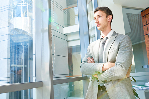 Portrait of confident young businessman standing by window with arms crossed looking away pensively, copy space