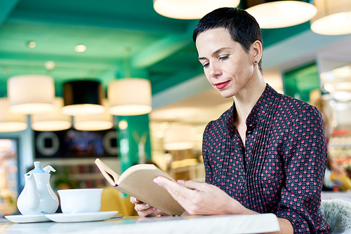 Portrait of modern adult woman smiling gently while reading book at table in cafe, copy space