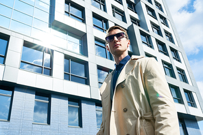 Low angle portrait of handsome young businessman wearing sunglasses and stylish trench coat standing outdoors looking away confidently against glass front of modern office building, copy space