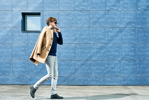 Full length portrait of handsome young man wearing sunglasses speaking by phone and smiling happily while walking in city street against grey concrete wall, copy space