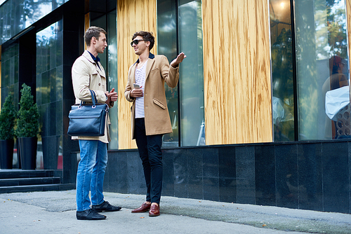 Full length portrait of two elegant young businessmen wearing autumn coats chatting outdoors in city streets, copy space