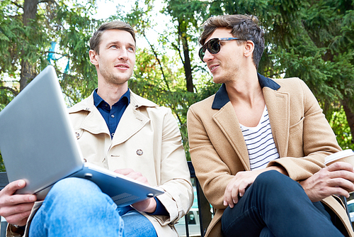 Low angle portrait of two handsome young men chatting and using laptop outdoors sitting on bench in park