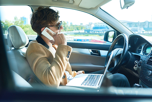 Side view portrait of successful modern entrepreneur using laptop and speaking by smartphone while working in luxury car, copy space