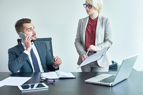 Attractive blond-haired manager showing financial figures to handsome male colleague while he talking to their boss on mobile phone, interior of open plan office on background