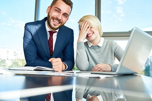 Group portrait of joyful young coworkers: handsome bearded man  with toothy smile while his female colleague covering her face with hand, panoramic office window on background