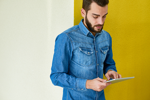 Young bearded businessman wearing denim shirt using digital tablet while standing against bright yellow wall of office lobby, waist-up portrait shot