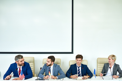 Row of confident politicians in formalwear sitting at table and listening to report prepared for international conference