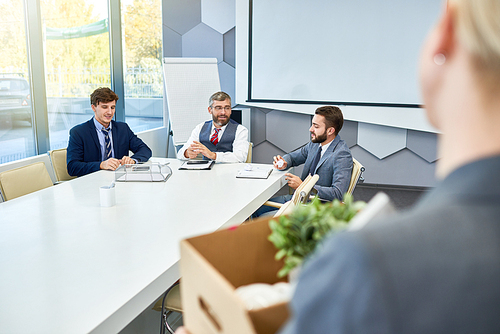 Group of hard-working white collar workers in classical suits sitting at boardroom table and analyzing results of accomplished work, unrecognizable female colleague holding cardboard box in hands