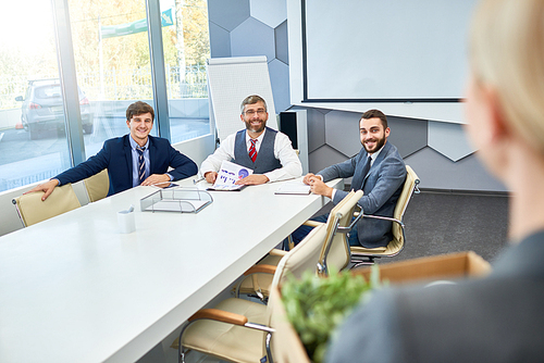 Rear view of unrecognizable young woman holding box of personal belongings coming to business interview in board room, focus on three cheerful business people greeting job interview candidate