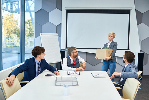 Group of white collar workers distracted from project discussion and looking at pretty middle-aged colleague with cardboard box in hands, interior of spacious boardroom on background