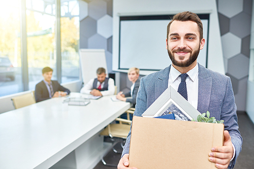 Waist-up portrait of handsome bearded manager in classical suit looking away with wide smile while holding cardboard box with belongings in hands, interior of spacious boardroom on background