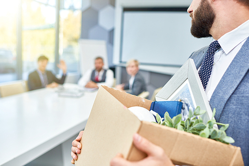 Unrecognizable man holding box of personal belongings looking back at several business people who fired him