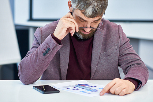 Waist-up portrait of concentrated middle-aged manager sitting at office desk and analyzing statistic data, blurred background