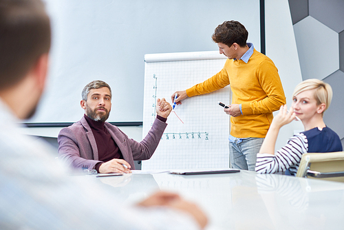 Handsome young businessman standing at marker board and drawing productivity graph while his colleagues sitting in boardroom and listening to him