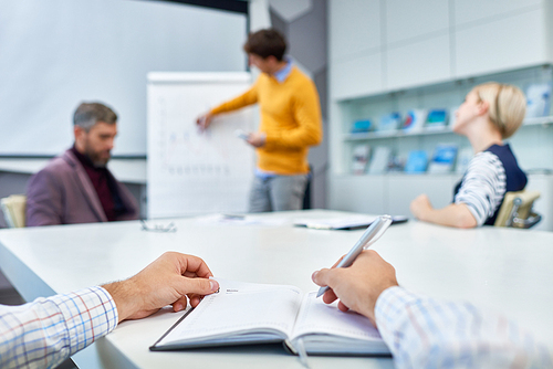 Close-up shot of unrecognizable manager taking necessary notes while sitting at boardroom table during working meeting, focus on foreground