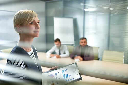 Portrait of ambitious young woman standing in office looking away hopefully and smiling with three people at meeting table in background, copy space