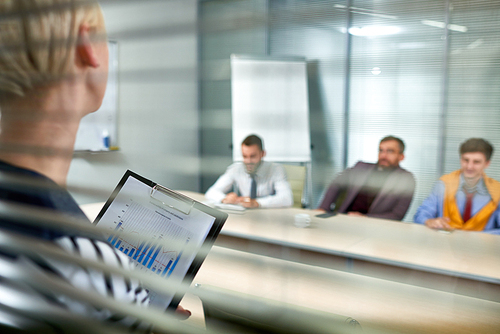 Unrecognizable manager with clipboard in hands presenting results of accomplished work to male colleagues while having working meeting at boardroom, view through glass wall