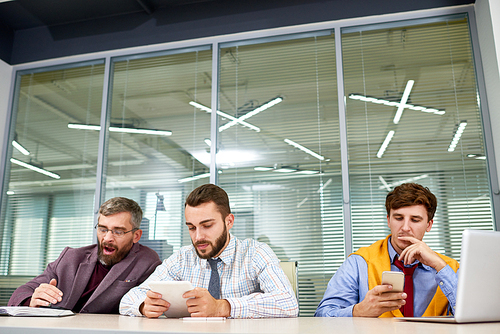 Three bored business  people waiting for meeting and using gadgets sitting in row at table in board room