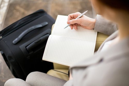 High angle close up of unrecognizable young businesswoman writing in notebook while waiting for business travel with suitcases
