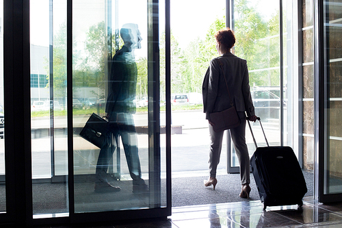 Back view silhouette of young businesswoman with suitcase walking through sliding glass doors of hotel, copy space