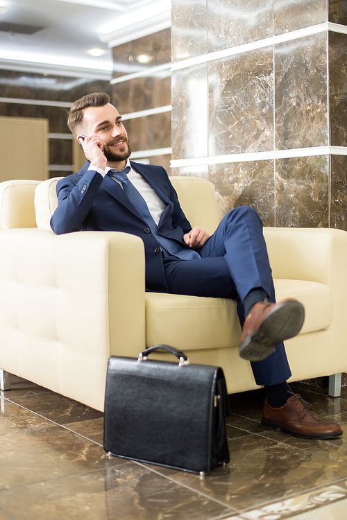 Full length portrait of handsome bearded businessman speaking by phone and smiling while waiting in hotel lobby sitting in leather armchair