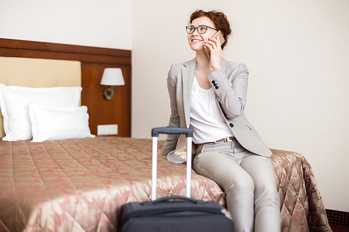 Portrait of happy young woman speaking by phone and smiling while sitting in hotel room on bed arriving for business travel, copy space