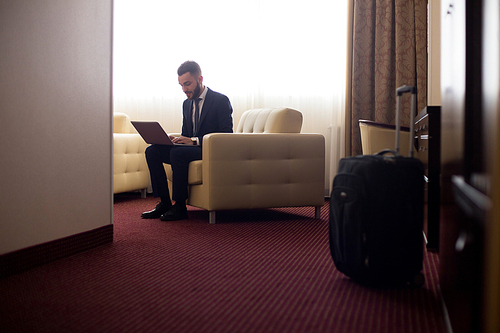 Full length portrait of handsome young businessman using laptop in hotel room, sitting in armchair with suitcase in foreground, copy space