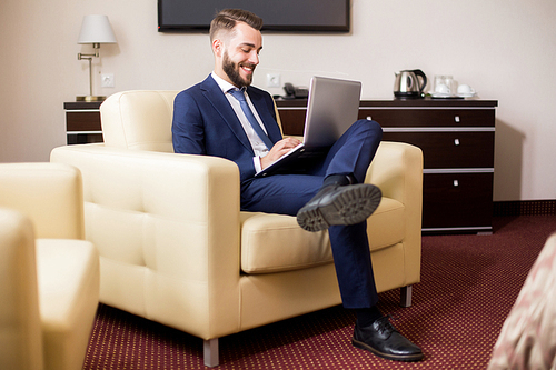 Full length portrait of handsome young businessman using laptop in hotel room and smiling while relaxing casually in armchair