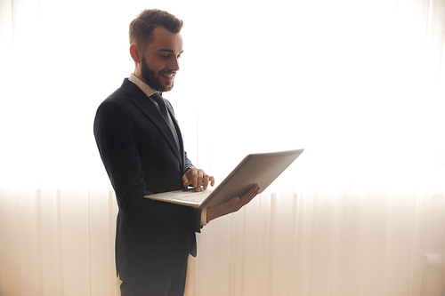 Side view of successful bearded businessman using laptop and smiling standing against background of window curtains in hotel room, copy space