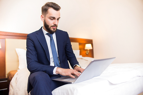 Portrait of handsome bearded businessman using laptop sitting on bed in comfortable hotel room during business travel, copy space