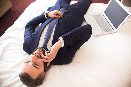 Above view of happy handsome businessman speaking by phone and smiling laying on hotel bed next to laptop, enjoying business travel, copy space