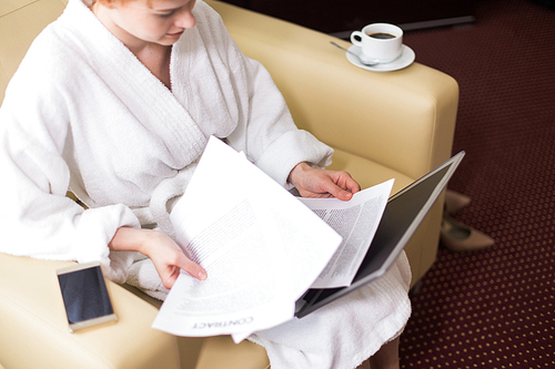 High angle portrait of young businesswoman dressed in bathrobe working with laptop and documents in hotel room during business travel, copy space