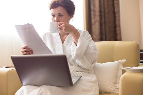 Portrait of young businesswoman dressed in bathrobe working with laptop and reading documents in hotel room enjoying business travel, copy space