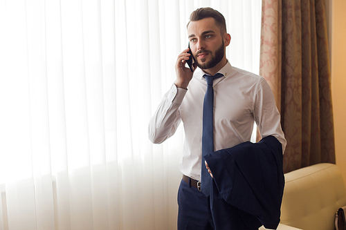 Portrait of handsome bearded businessman speaking by phone standing by window in hotel room enjoying business travel