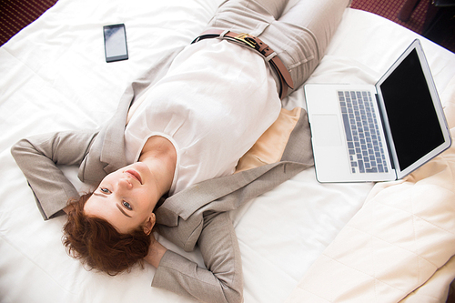 Directly above portrait of smiling young businesswoman laying on hotel bed next to laptop relaxing and enjoying business travel