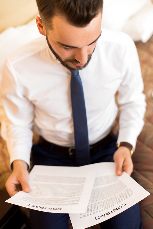 High angle portrait of young businessman working with documents sitting on hotel bed during business travel